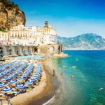 sandy beach with blue and white beach umbrellas with a white cliffside town in the background