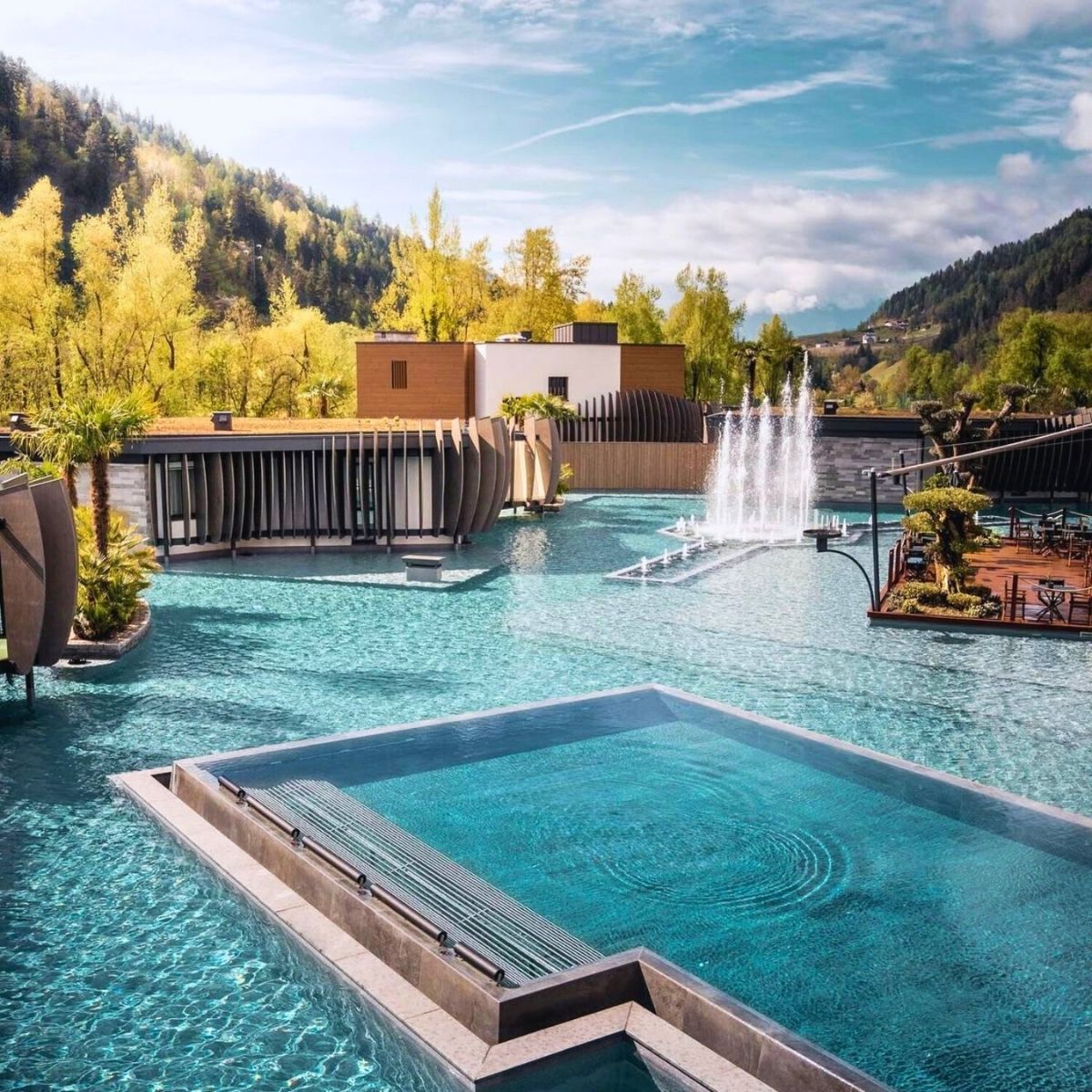 large swimming pool with a fountain and tree-lined mountains in the background
